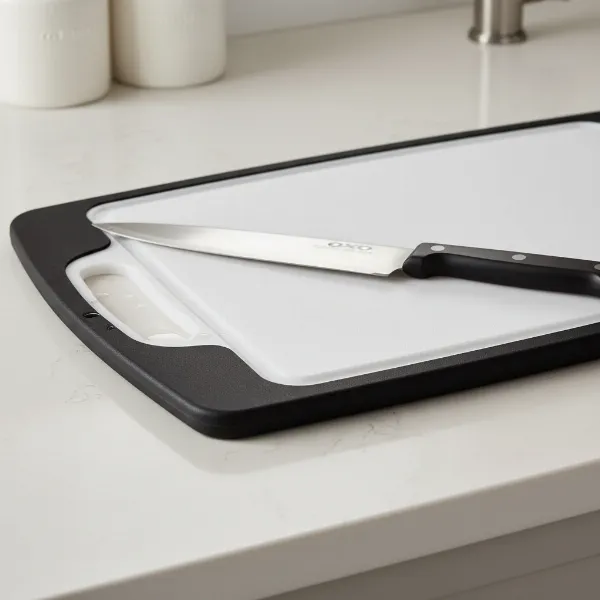 A close-up shot of the OXO Good Grips Utility Board, clearly showing its rubberized non-slip edges and the juice groove on one side, with a chef's knife resting on it.